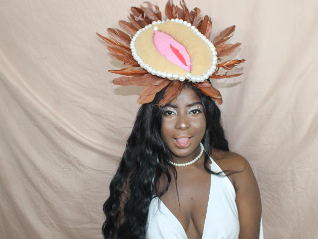 EmmaBlosson with dark skin, long black hair, white halter top, pearl necklace, sticking out tongue, wearing brown feather and pink heart hat, showing big tits.