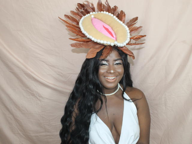 EmmaBlosson with dark skin, long wavy black hair, wearing a white halter top showing big tits, pearl necklace, and a brown feathered hat with a pink and white oval.