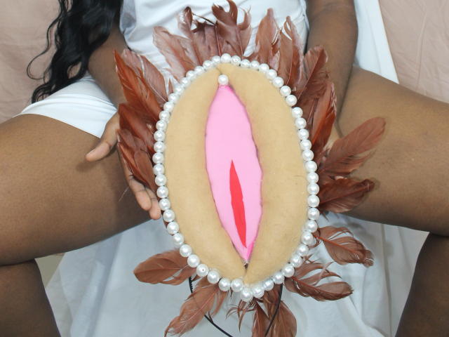 EmmaBlosson's hands hold a fake vagina made of pink and beige fabric, surrounded by brown feathers and white pearls, with her dark-skinned thighs visible.