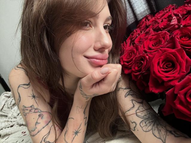SensualMila with long brown hair, smiling softly, resting chin on hand, showing tattoos on arms, next to a large bouquet of red roses, in a cozy, intimate setting.