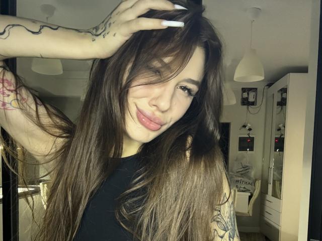 SensualMila with long brown hair, white nails, tattoos on arms, smiling, pink lips, black top, hand in hair, warm lighting, cozy room, slightly messy, sexy, confident.