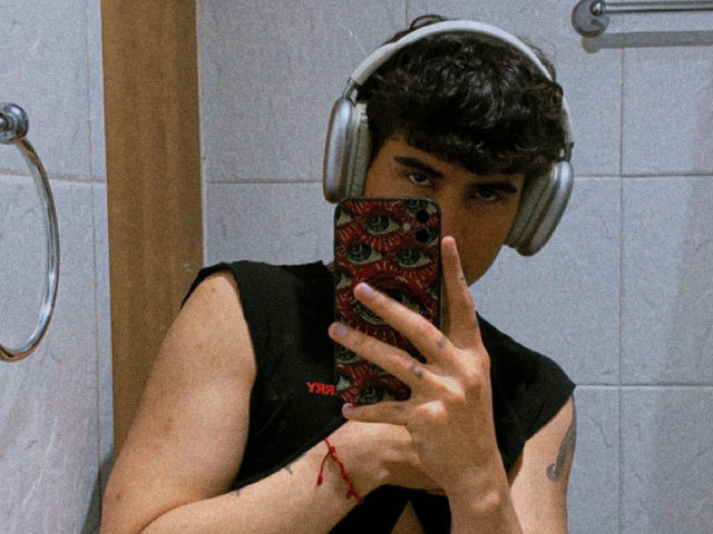 TobbyRamirez, wearing headphones, takes a mirror selfie with a phone featuring eye pattern cover, black sleeveless shirt, red bracelet, and tattoo on right arm.