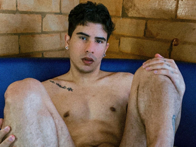 TobbyRamirez, shirtless, with tattoos, dark curly hair, and ear piercings, sitting with legs up, hands on knees, looking at camera, blue couch, brick wall background.