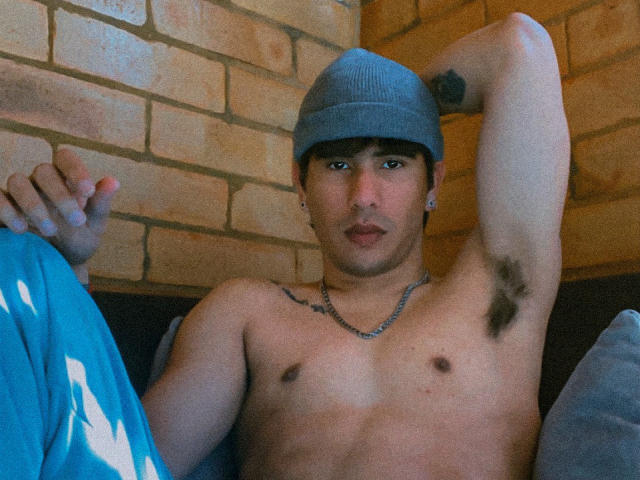 TobbyRamirez is shirtless, wearing a blue cap, and has armpit hair. He's sitting with one arm raised, a chain necklace, and a tattoo on his right shoulder.