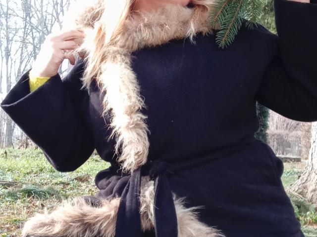 KelliRedhaer's black dress with fur trim, tied at the waist, showing fluffy fur on the chest and bottom, holding green pine leaves near her face.