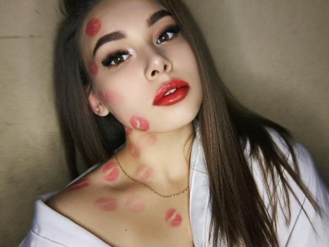 Niferttiti with red lipstick, long brown hair, and kiss marks on her face, neck, and chest, wearing a white shirt, looking at the camera with seductive eyes.