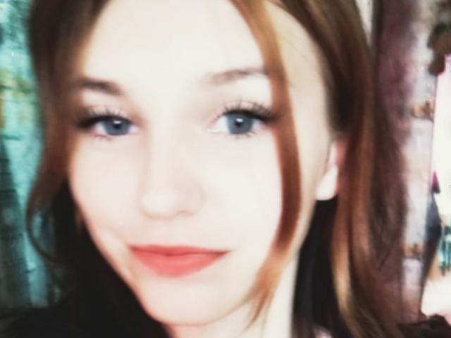 ShellyLili ha gli occhi azzurri, la pelle chiara e i capelli castani con una riga laterale, indossa un rossetto rosso e un eyeliner nero, e sorride dolcemente alla fotocamera..