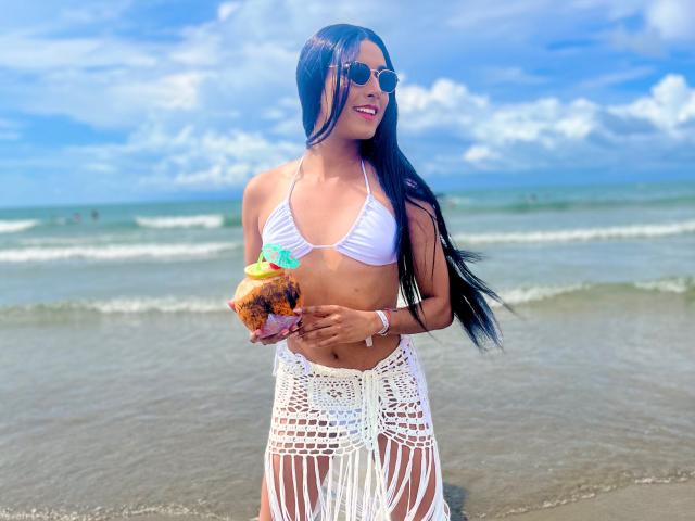 FallonRogers with long black hair, wearing a white bikini top and white crochet skirt, holding a coconut with a colorful straw, standing on a sandy beach, smiling.