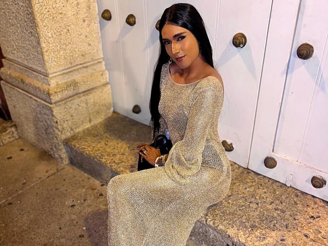 FallonRogers with long black hair, wearing a sparkly, off-shoulder, gold dress, sitting on a stone step, holding a black clutch, smiling, looking sexy, butt visible.