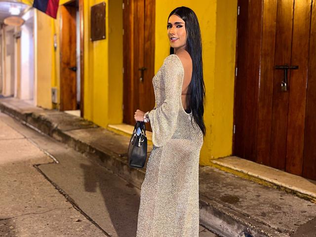 FallonRogers in a sparkly, sheer dress with long black hair, holding a black handbag, standing on a narrow street with yellow and brown doors. Her ass is visible.