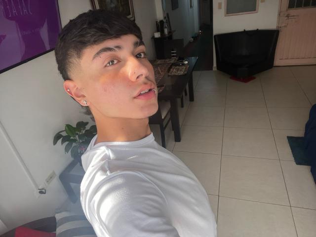 ImJuanda with short dark hair, a nose ring, and earrings, wearing a white shirt, looking over shoulder, sunlight on cheek, white-tiled floor, black chair in background.