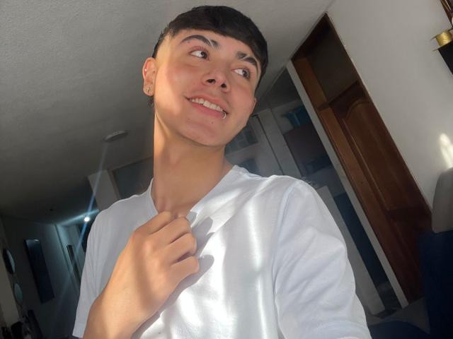 ImJuanda with short dark hair, white shirt, and light skin, smiling and holding shirt collar, sunlight on face, slightly tilted head, and brown eyes, in a sunlit room.