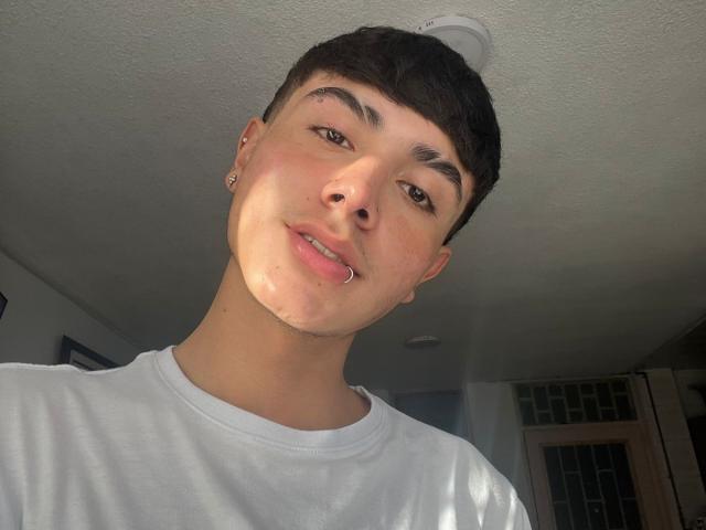 ImJuanda with short dark hair, brown eyes, and a nose ring, wearing a white shirt, has a slight smile, and is looking down at the camera. Sunlight on face.
