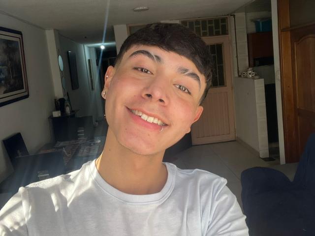 ImJuanda has a cute smile with short brown hair, wearing a white shirt, and small earrings, showing a happy, slightly dimpled face, standing in a sunlit room.