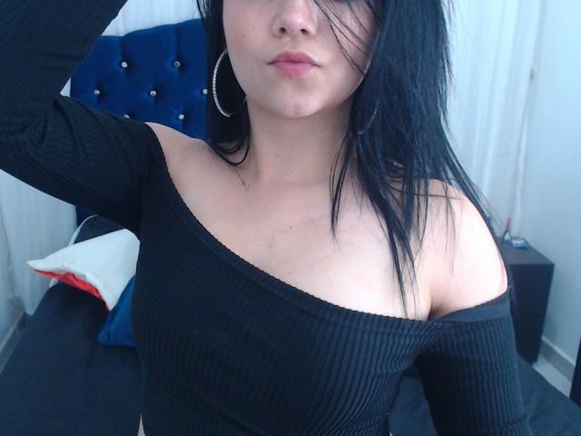 Kkatalina with black ribbed off-shoulder top, showing her tits, black hair, and hoop earrings, pouting with her hand in her hair. Blue tufted headboard in background.