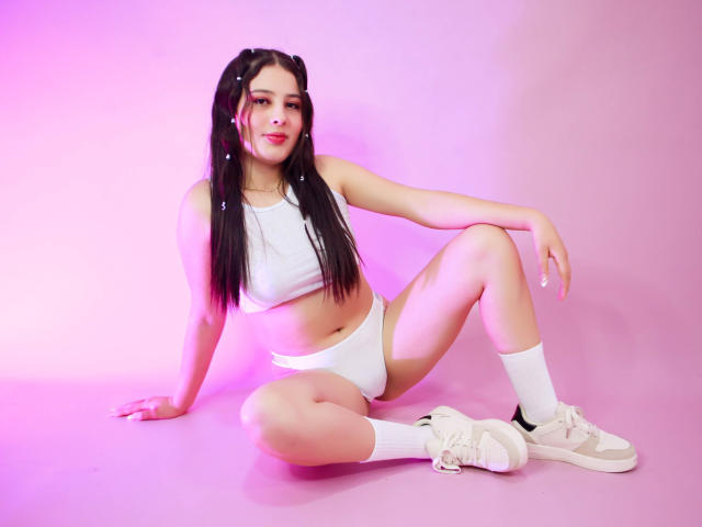KathyStone with long black braids, white crop top, white panties, white socks, white sneakers, sitting on pink floor, legs spread, arms resting, smiling, pink lighting.