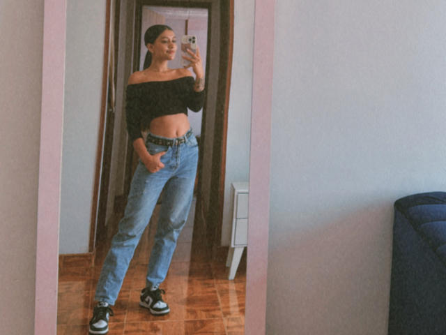 ThaliaAres with a black off-shoulder crop top, blue high-waisted jeans, black and white sneakers, standing in front of a mirror, showing her ass and midriff.