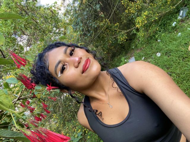 SophieSonyas with curly black hair, wearing a black tank top, red lipstick, and eyeliner, smiling at the camera, surrounded by red flowers and greenery.