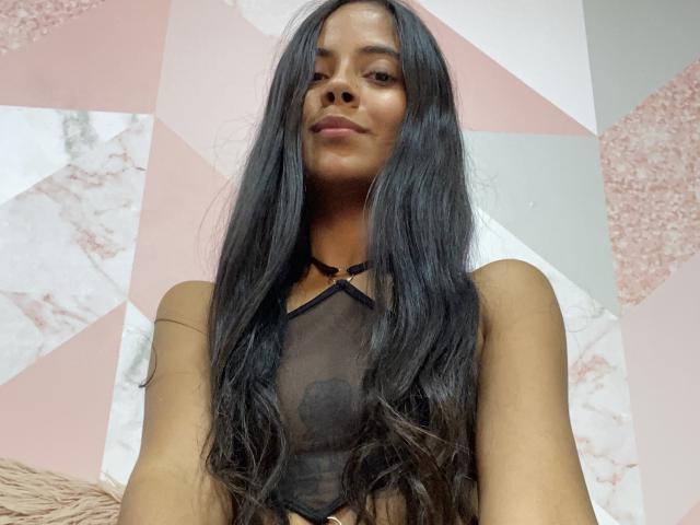 TamaraPeach, com longos cabelos pretos, vestindo uma blusa preta transparente com um pequeno coração preto, mostrando seus pequenos mamilos pretos. Ela tem um leve sorriso e pele escura..