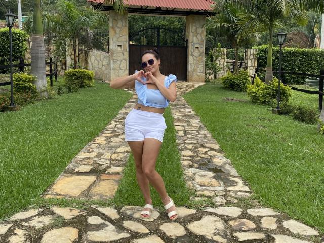 GalaMalkova standing on a stone path, wearing a blue tied-up crop top, white shorts, white sandals, and sunglasses, with her hand near her mouth, showing her ass.
