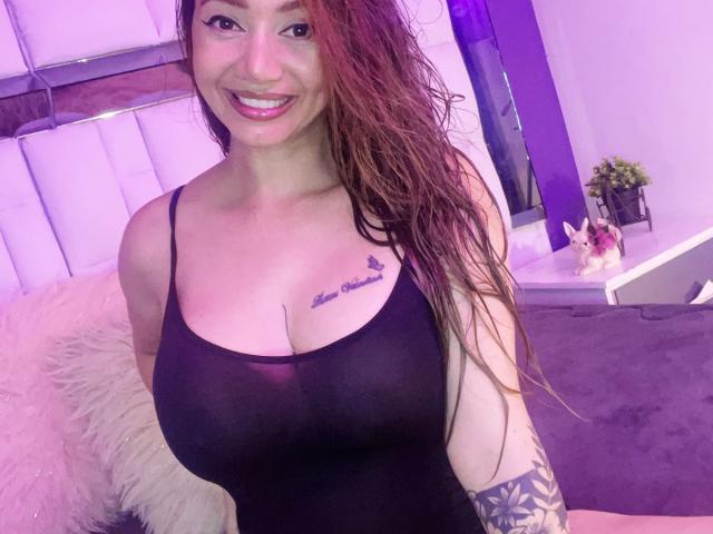 SophieLombardo with wet red hair, smiling, wearing a sheer black top that shows her big tits and tattoo above her cleavage. She has a tattoo sleeve on her right arm.