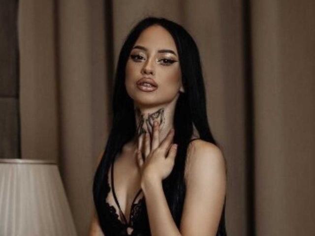 MassimoMillen with long black hair, wearing a black lace top, has tattoos on her neck and hand, looking sexy with pouty lips, standing by a lamp.