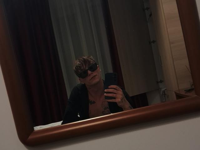 MassimoMillen with short brown hair, black sunglasses, black shirt, chest tattoos, holding phone in mirror. Mirror has wooden frame, dimly lit room. MassimoMillen's phone is black.