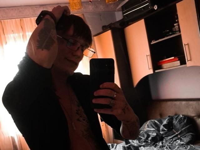 MassimoMillen taking a mirror selfie, wearing glasses, black shirt, exposing chest tattoos, holding phone, right arm raised, showing tattoo, bed with rumpled blue sheets in the background.