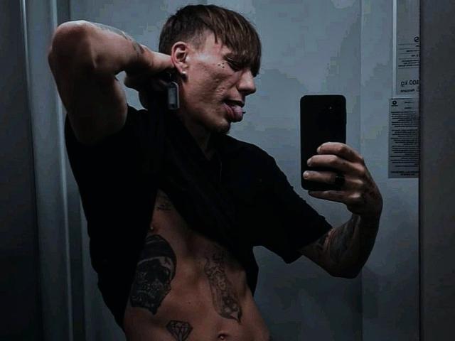 MassimoMillen, shirtless with tattoos, arms raised, taking a mirror selfie with a phone, tongue out, in a dimly lit, gray-walled elevator.