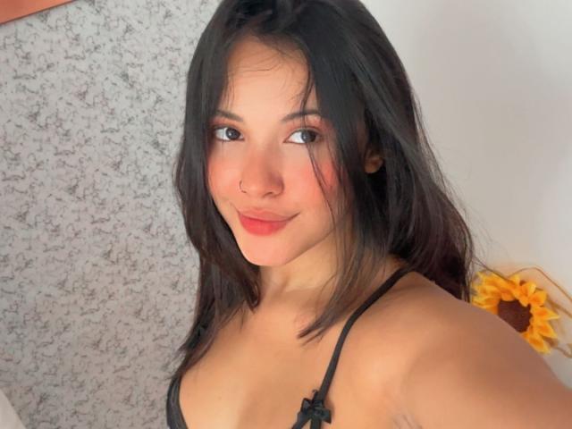 ArianaSmith69, com cabelo castanho escuro, sombra rosa, piercing no nariz, sutiã de renda preto, sorrindo para a câmera, girassol ao fundo, pele clara, seios médios, selfie casual..