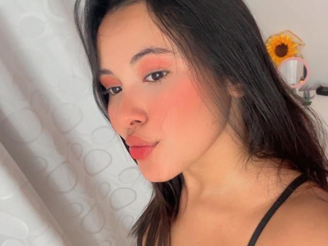 ArianaSmith69 with pink eyeshadow, rosy cheeks, and red lips, black hair, black spaghetti strap, subtle smile, soft light, white curtain with circular pattern in background.