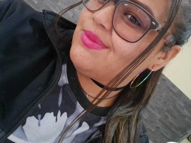MelannieCarter with pink lipstick, glasses, and a black choker, wearing a Batman shirt, black jacket, and colorful hoop earrings, smiling at the camera.