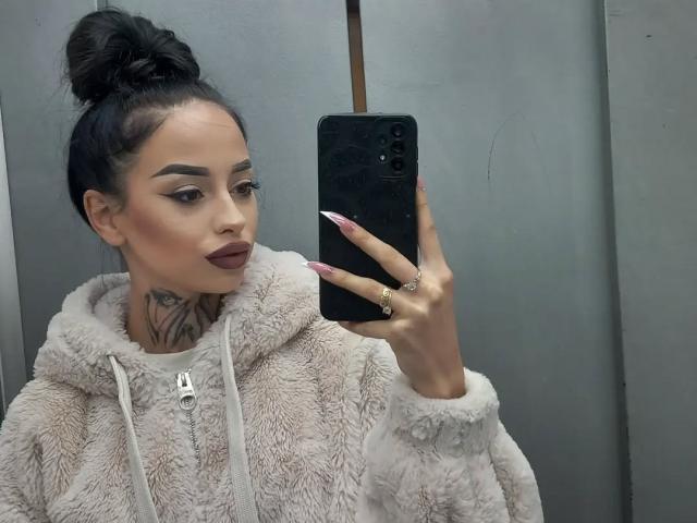 MassimoMillen with dark hair in a bun, wearing a fluffy beige hoodie, holding a phone, showing pink nails, and a tattoo on the neck, with maroon lipstick.