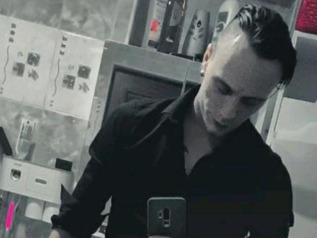 MassimoMillen with a side-swept haircut, wearing a black shirt, looking down at a smartphone in a dimly lit room, holding a pink object in his left hand.