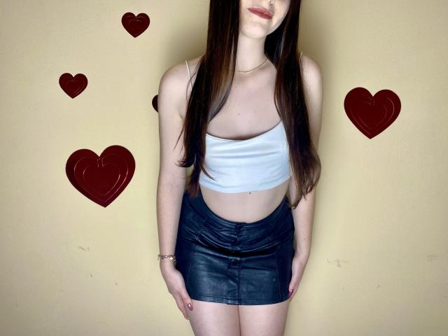 Valissia with long brown hair, white crop top, showing small tits, and black leather mini-skirt, standing, hands by sides, yellow wall with red heart stickers.