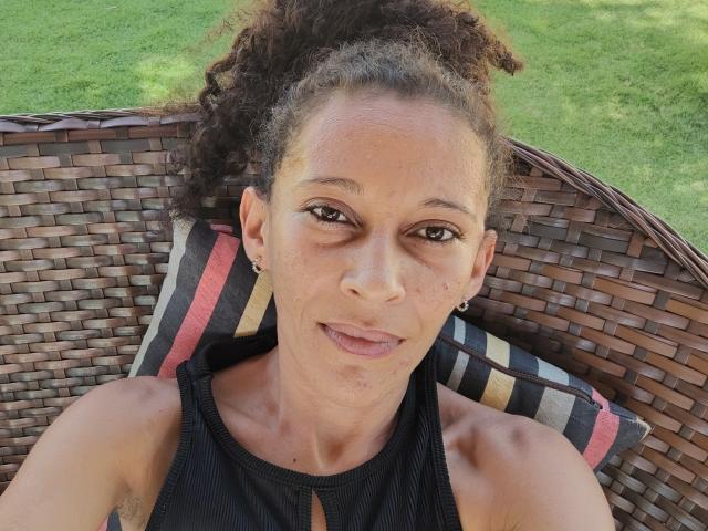 ManuZinhah with curly brown hair, tan skin, small earrings, black sleeveless top, sitting on wicker chair with striped pillow, smiling at camera, visible small tattoo on right shoulder.