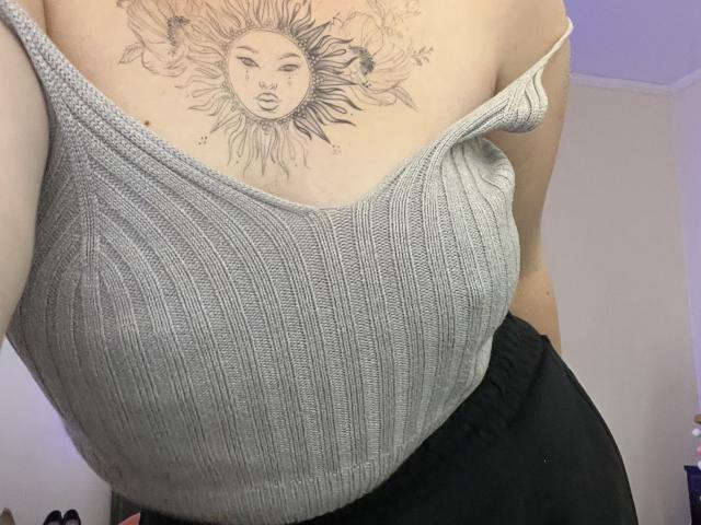 DiLaria's grey ribbed tank top shows off her big tits, with a sun tattoo on her chest. She's wearing black pants, and the tattoo has sun rays and flowers.