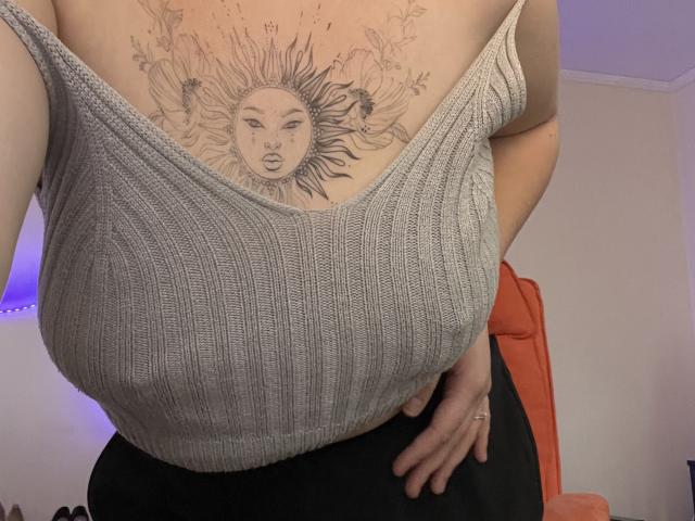 DiLaria's large tits in a ribbed, gray tank top, showing a sun tattoo with flowers on her chest. She's wearing black pants, hand on hip. Visible orange chair.