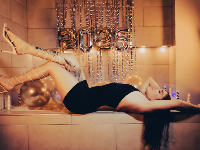 AndyDupont, allongée sur le dos, portant une robe noire, des talons hauts et des bottes à lacets, les jambes levées, montrant sa partie intime, dans une pièce décorée avec des ballons dorés et des éléments décoratifs "2023"..