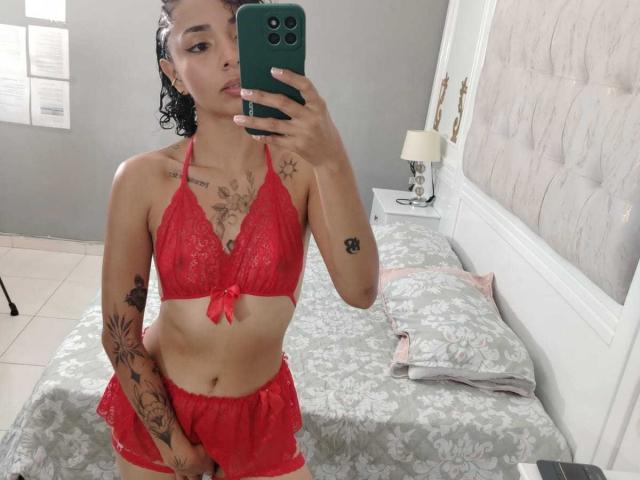 SilvanaXOwen in red lace bra and shorts, holding phone, showing tattoos, with curly black hair, light skin, and small tits, standing in front of a bed with grey bedding.