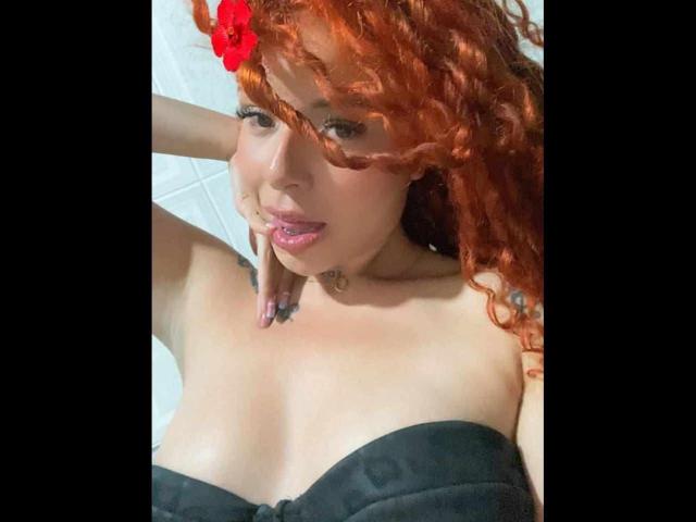 TammyyRoss, avec ses cheveux roux bouclés, portant un haut noir sans bretelles, montrant ses seins, avec son doigt dans la bouche, et une fleur rouge dans ses cheveux..