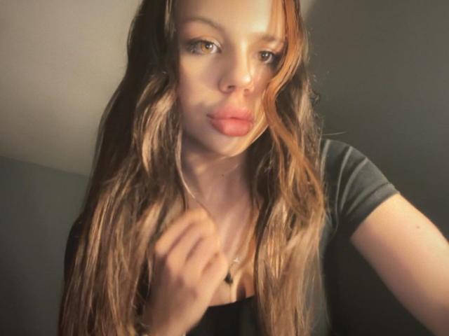 DeineLexy-hot with long brown wavy hair, wearing a black shirt, slightly pouting lips, holding her shirt, looking at the camera, with a subtle, sexy, confident expression.