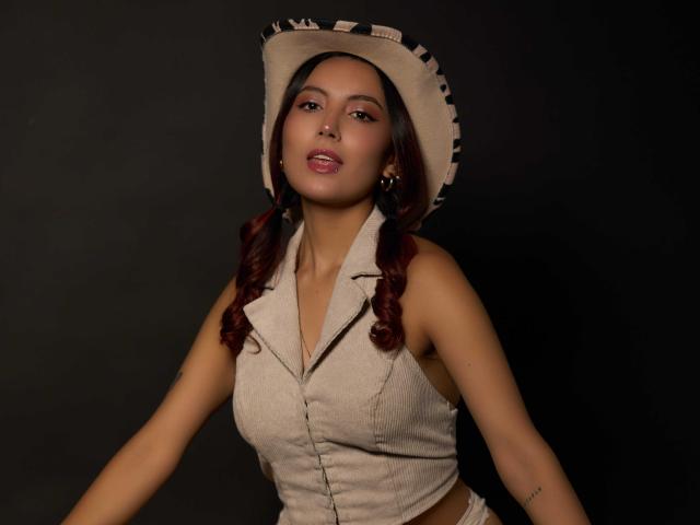 CarlaFranco with dark brown hair in pigtails, wearing a beige cowboy hat and sleeveless button-up vest, showing her tits against a dark background.