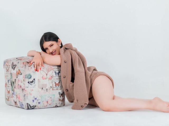 Emma Lombardi, met zwart haar, liggend op haar zij, gekleed in een bruine wollen jurk met knopen, leunend tegen een gestreept kubus, waarbij haar kale, bleke en ronde billen zichtbaar zijn..