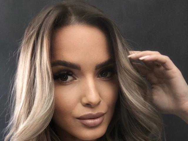 AnnetteBriane has blonde, wavy hair, brown eyes, and full, nude-colored lips. She's touching her hair with a hand, showing a subtle, confident smile. Her skin is tan.