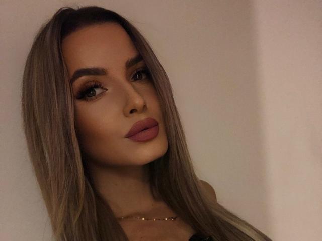 AnnetteBriane has long brown hair, full eyebrows, and pouty pink lips. She's wearing a gold necklace and looking at the camera with a serious expression.