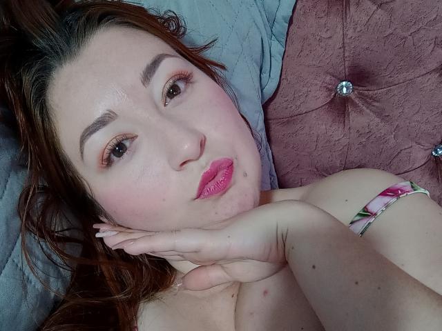 ReginaLov with pink lipstick, brown eyes, and brown hair, pouting slightly, hand under chin, showing her tits, on a pink, tufted, velvet pillow.