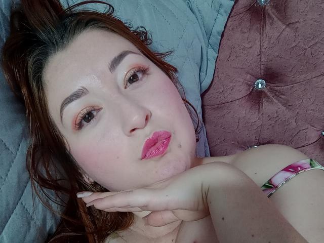 ReginaLov with pink lipstick, brown eyes, and wavy brown hair, pouting, resting her chin on her hand, showing her bare shoulder with a pink floral bracelet.