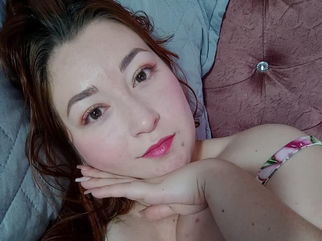 ReginaLov with fair skin, brown eyes, pink lipstick, and red hair, lying on a bed, her hand under her chin, showing her bare shoulder and floral strap.