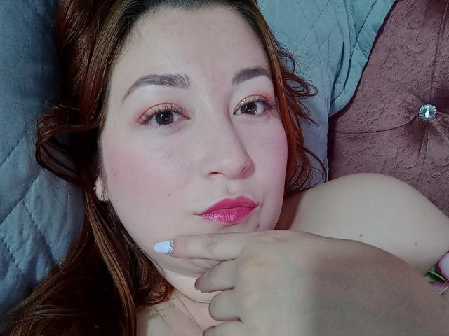 ReginaLov has fair skin, pink lipstick, and brown hair, lying down with a finger near her chin. Her pink-tinted eyelids match her glossy lips. Boobs partially visible.